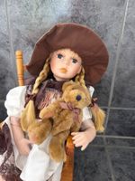Porzellanpuppe mit Teddy auf Stuhl sitzend